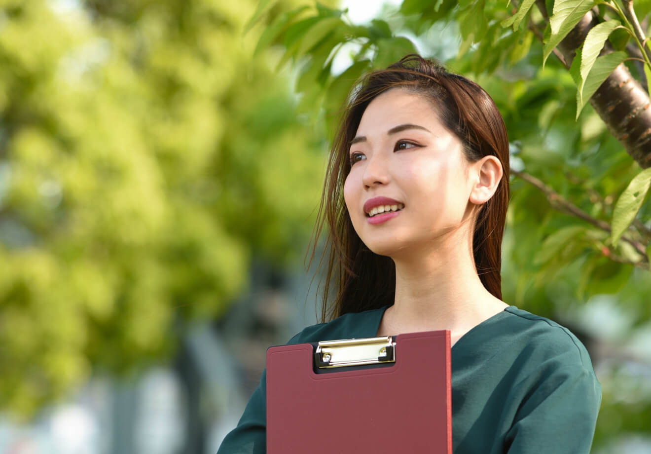 屋外の緑茂る木の前にて、バインダーを抱えながら立つ女性